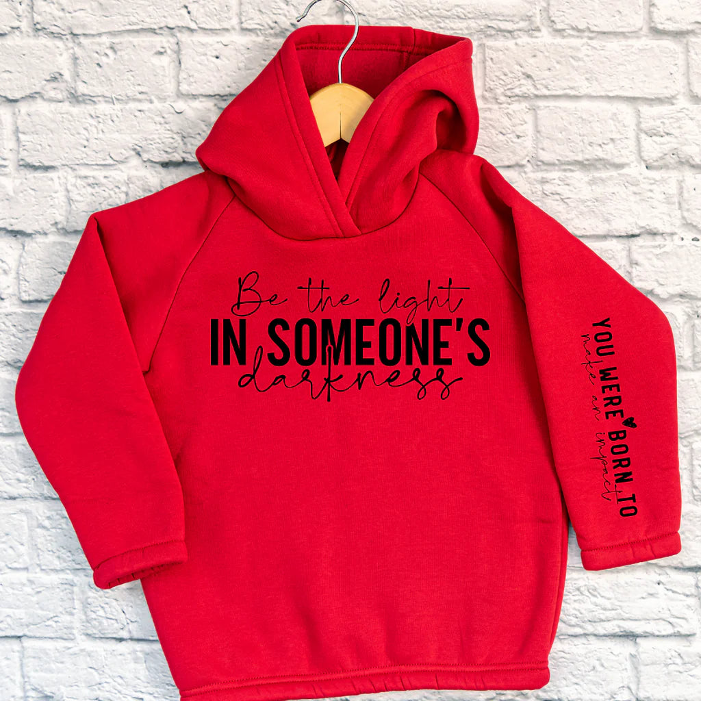 Red cotton blend hoodie with the inspirational message "Be the Light in Someone's Darkness" on the chest and "You Were Born to Make an Impact" on the sleeve, hanging up on a brick wall.