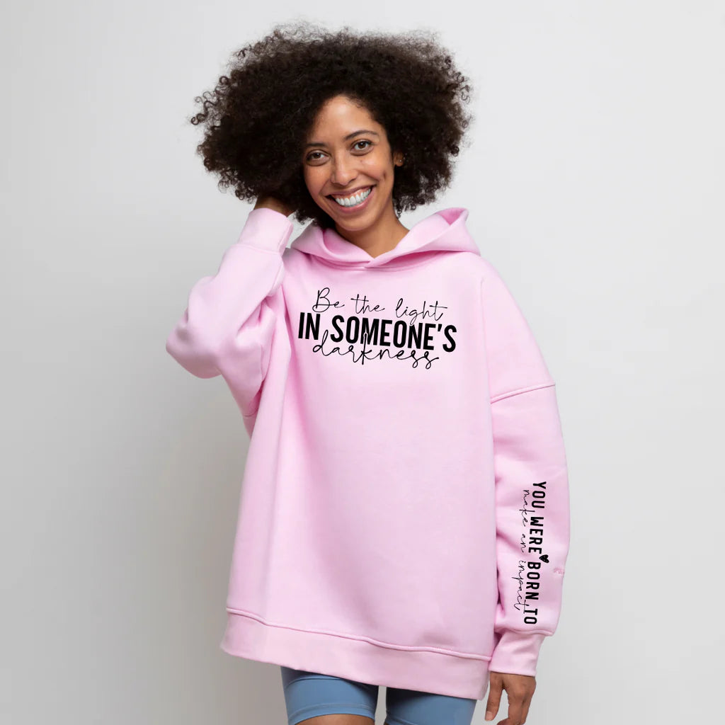 African American lady wearing a pink cotton blend hoodie with the inspirational message "Be the Light in Someone's Darkness" on the chest and "You Were Born to Make an Impact" on the sleeve, standing against a plain white background.