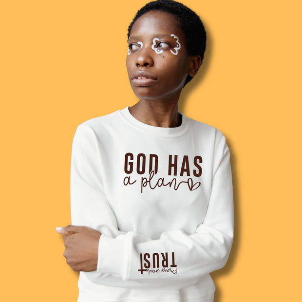 African American gril wearing a White cotton blend hoodie with the phrase "God Has a Plan" in bold brown lettering on the chest and "Pray, Wait, Trust" on the sleeve, her arms are crossed, and she has face painting around her eyes she is displayed against a light neutral background.